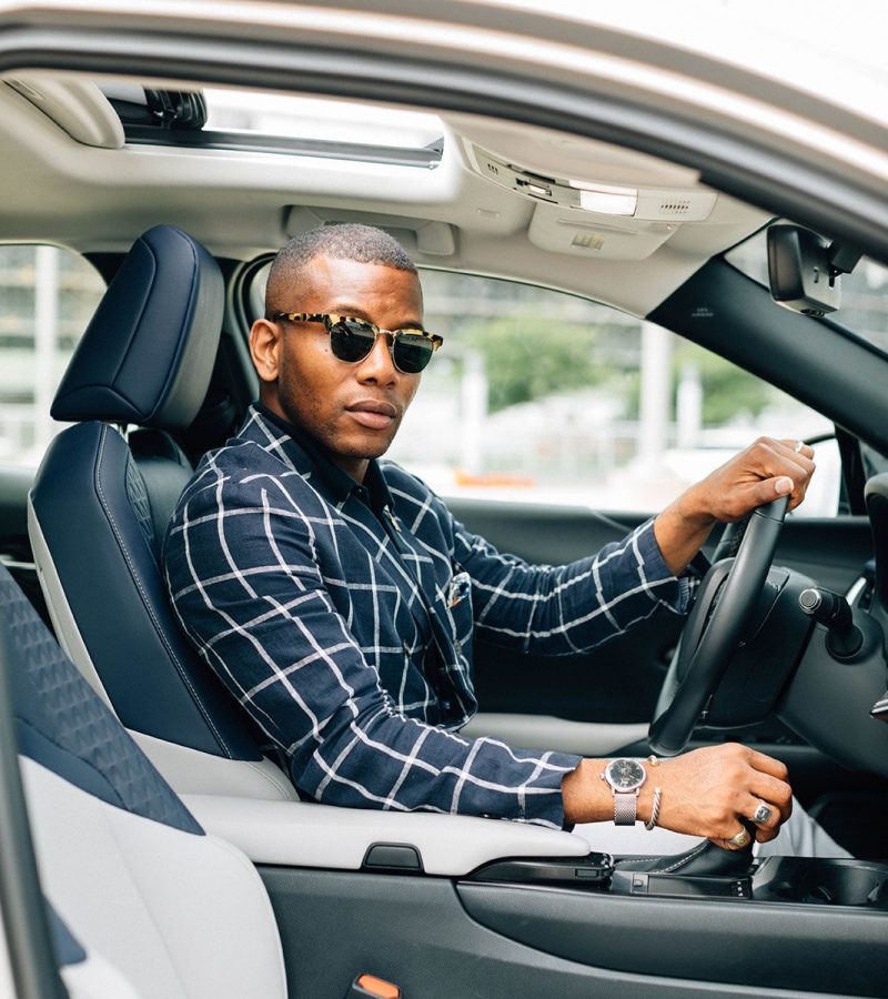 An image of a stylish man in the driver's seat of a car, showcasing a coordinated ensemble that includes a windowpane check shirt, tortoiseshell sunglasses, and a silver-tone mesh watch - The Curated Table