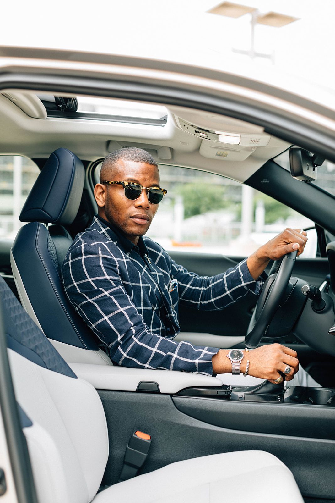 An image of a stylish man in the driver's seat of a car, showcasing a coordinated ensemble that includes a windowpane check shirt, tortoiseshell sunglasses, and a silver-tone mesh watch - The Curated Table