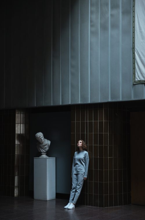 A woman leaning against a dark wall in an art gallery next to a classical style bust statue